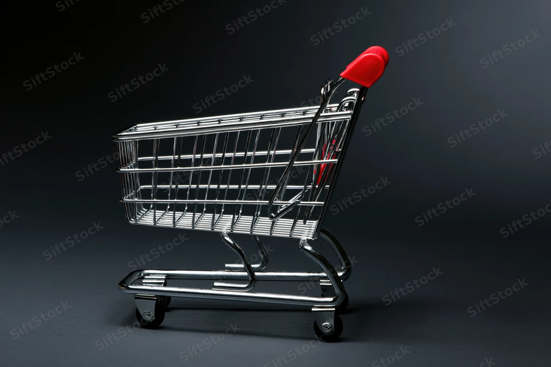 Small empty metal shopping cart with red plastic handle on dark studio background