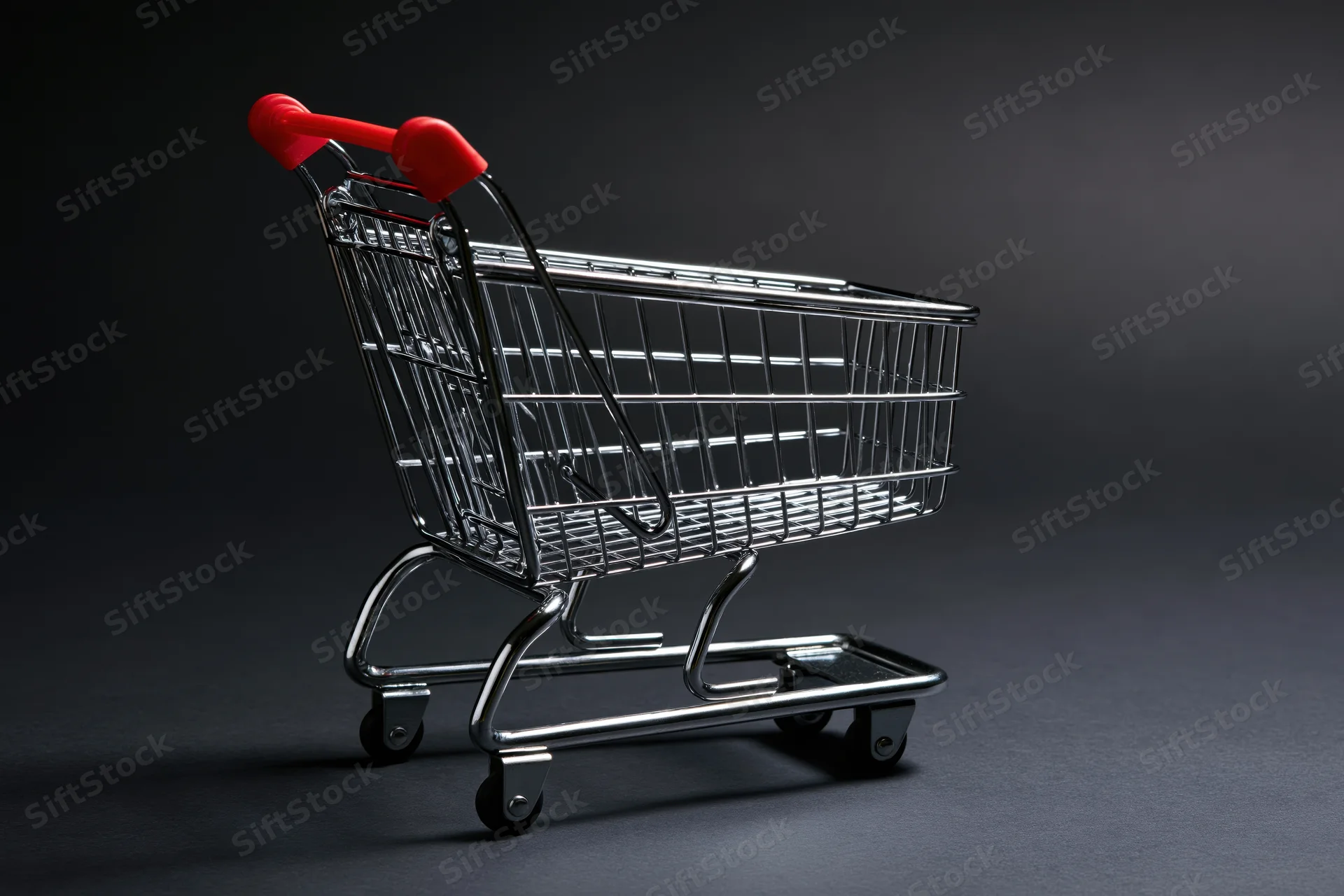 Small empty metal shopping cart with red plastic handle on dark studio background