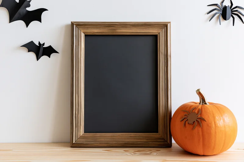 Small orange pumpkin and blank dark chalkboard in wooden frame on table with halloween decorations on wall