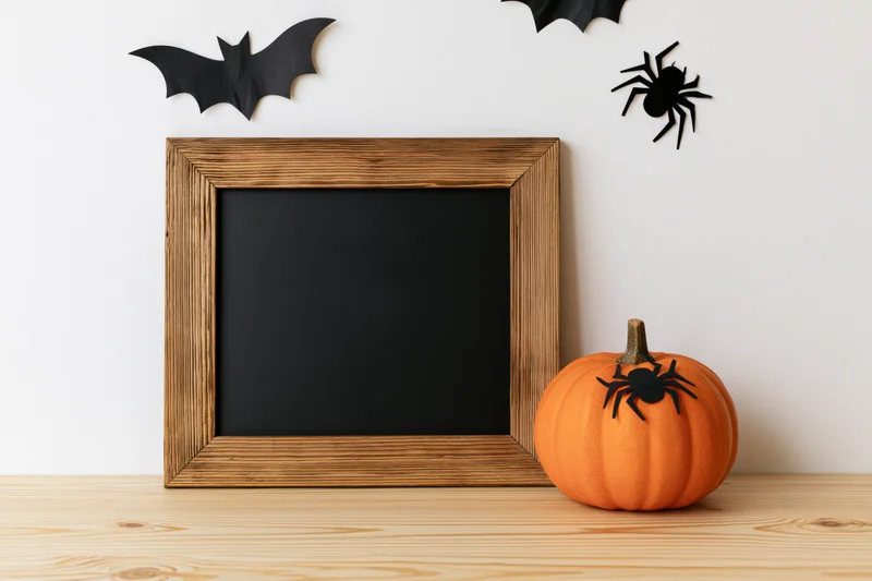 Small orange pumpkin with black spider decoration beside a blank dark chalkboard in a wooden frame on a wooden table with bats and spider on the wall above