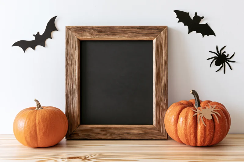 Two pumpkins flanking a blank chalkboard frame on a wooden table with halloween decorations on the wall