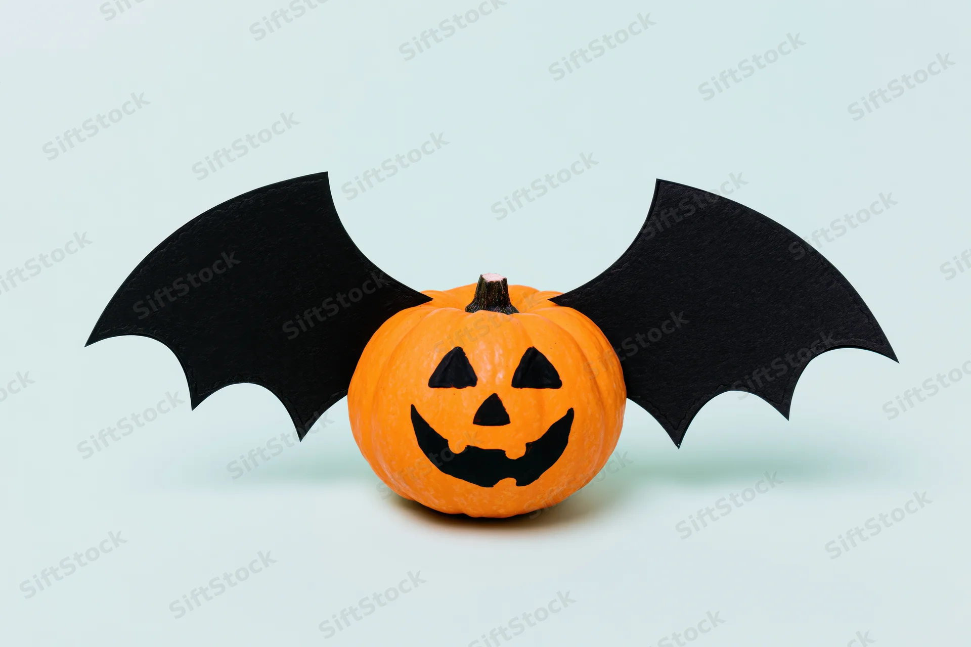 Small orange pumpkin with painted jack-o-lantern face and paper bat wings on light background