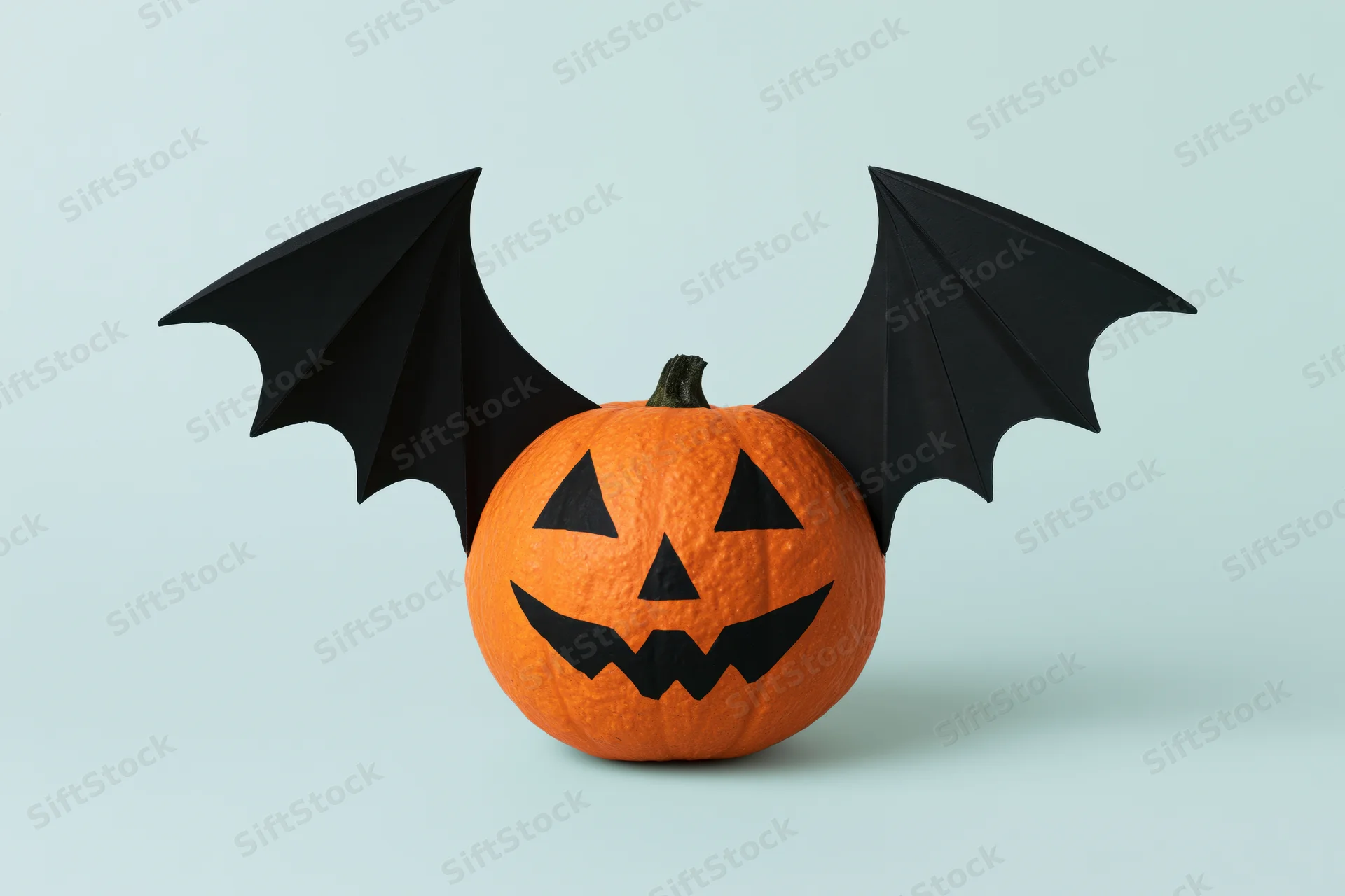 Small orange pumpkin with painted jack-o-lantern face and black bat wings on light blue background