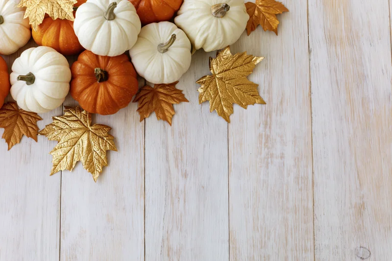 Small white and orange pumpkins with gold leaves arranged on a white wooden surface