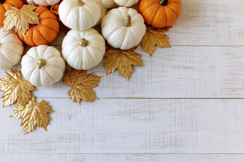 Small white and orange pumpkins with gold leaves arranged on a whitewashed wooden table