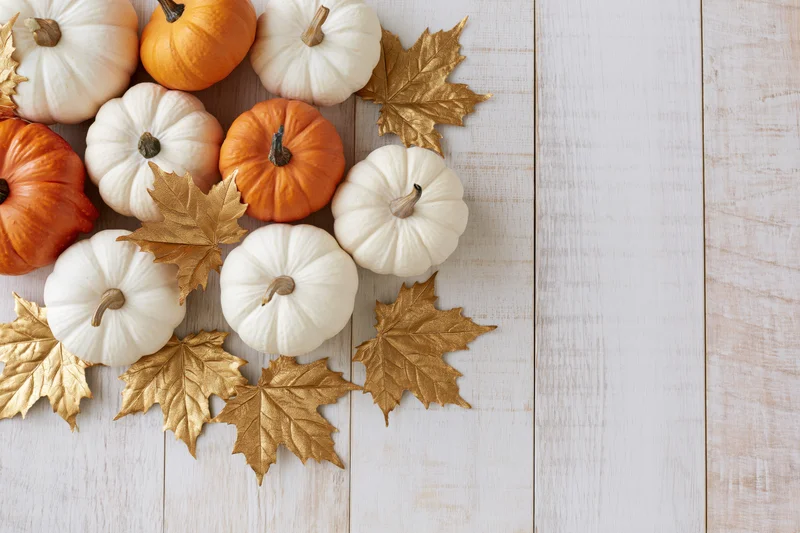 Small white and orange pumpkins with gold leaves arranged on a white wooden table