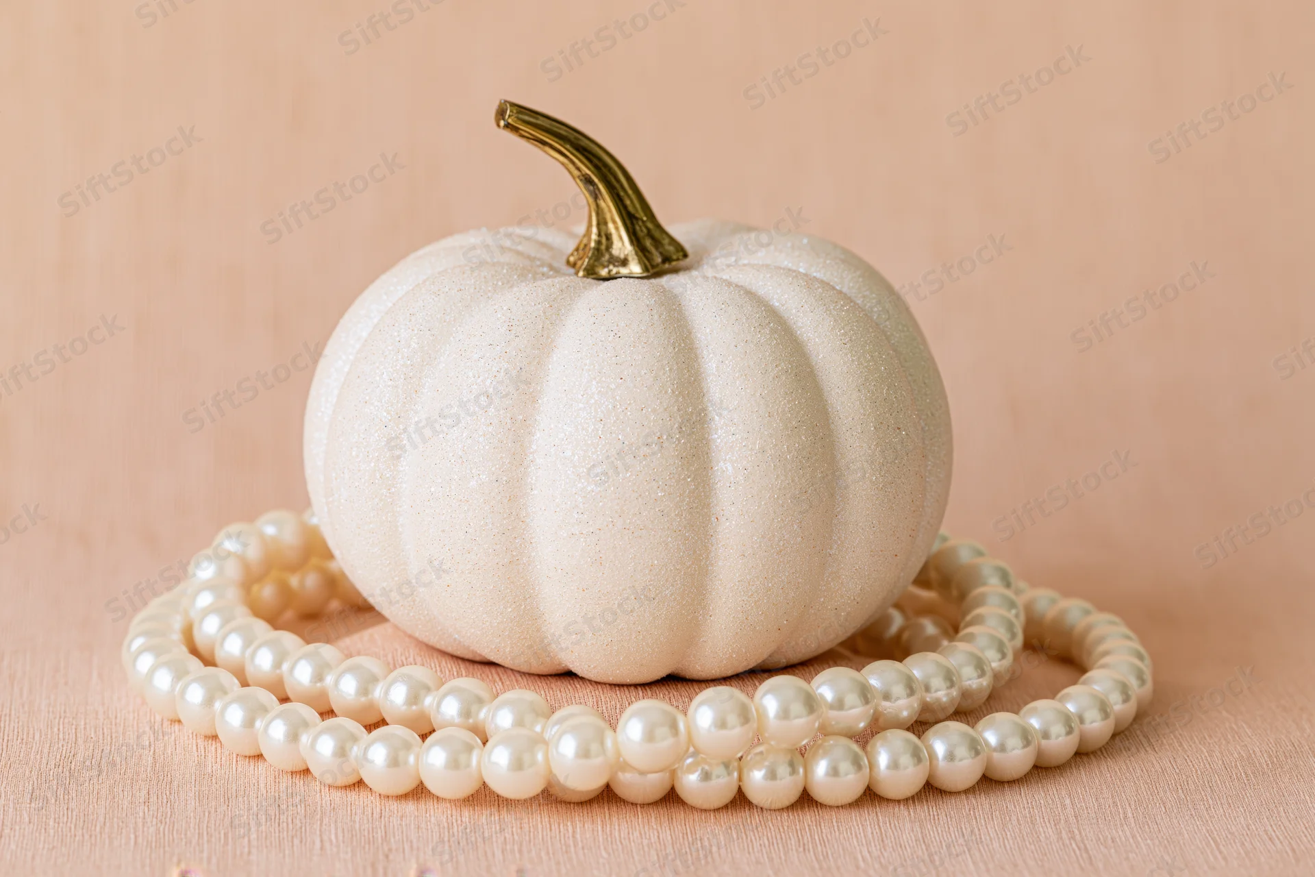 Small white decorative pumpkin with gold stem resting on a pearl necklace on a beige fabric surface