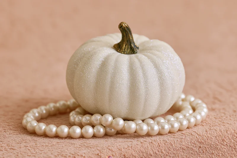Small white decorative pumpkin with glittery surface and gold stem resting on a pearl necklace atop a beige textured fabric