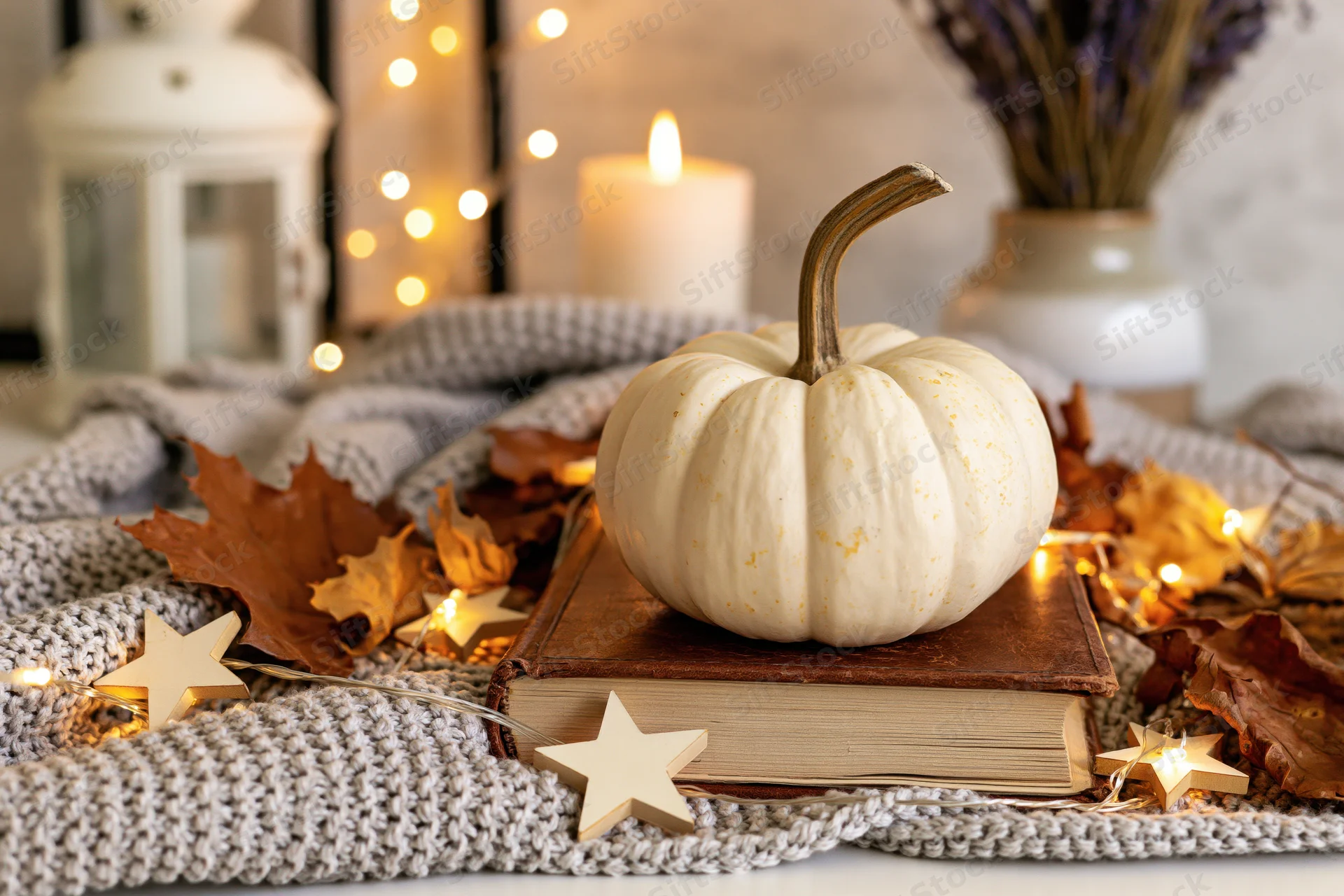 White pumpkin resting on a worn leather book surrounded by autumn leaves and fairy lights on a knitted blanket