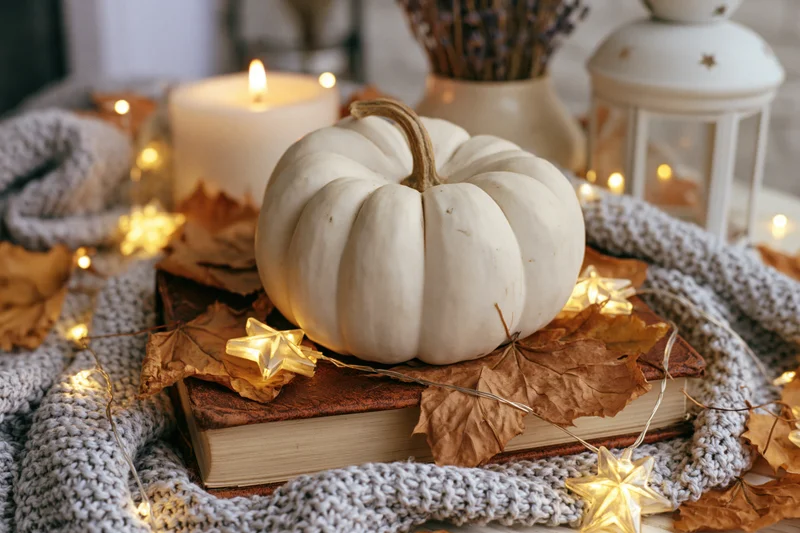 White pumpkin resting on a worn leather book surrounded by dried leaves and fairy lights on a knit blanket