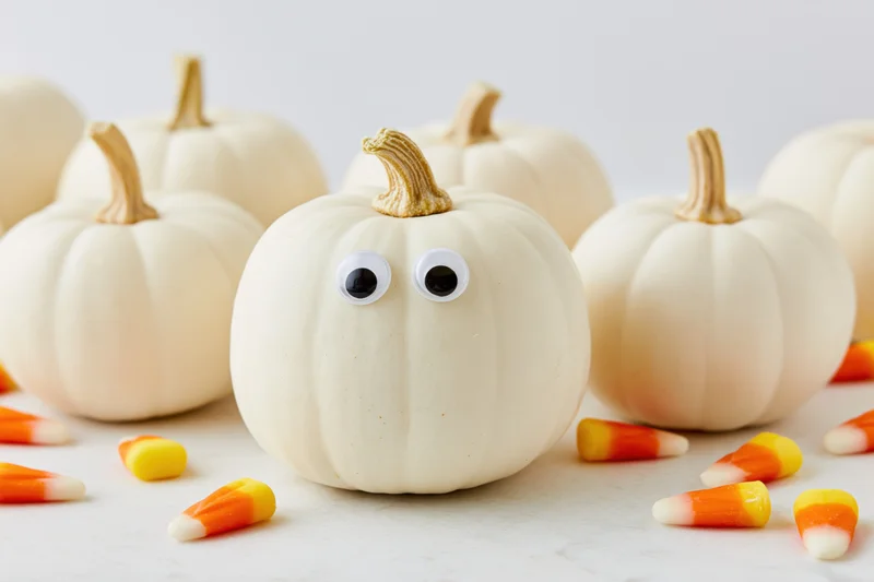 Small white pumpkins with googly eyes arranged on a white surface with candy corn scattered around