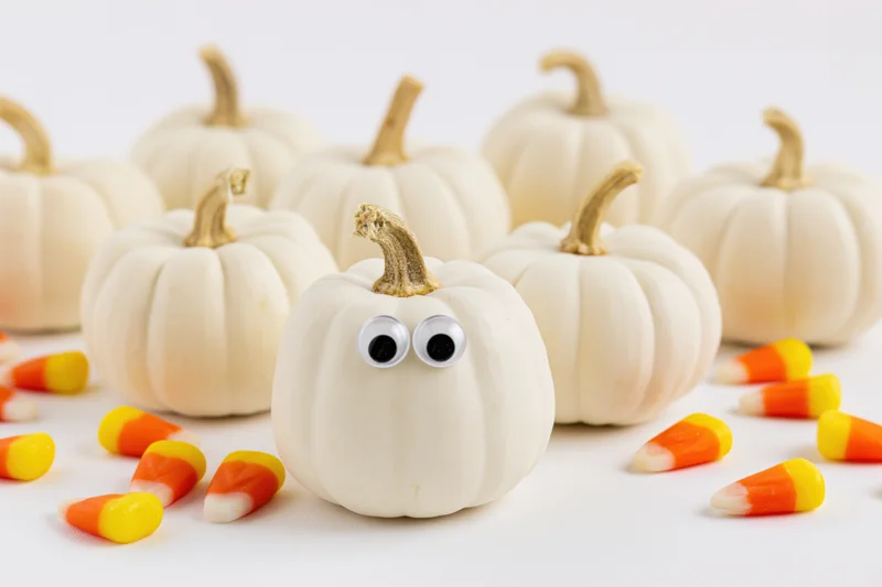 Small white pumpkins with googly eyes arranged on a white surface with candy corn scattered around