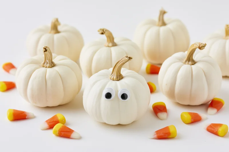 Small white pumpkins with googly eyes and candy corn scattered on a white surface