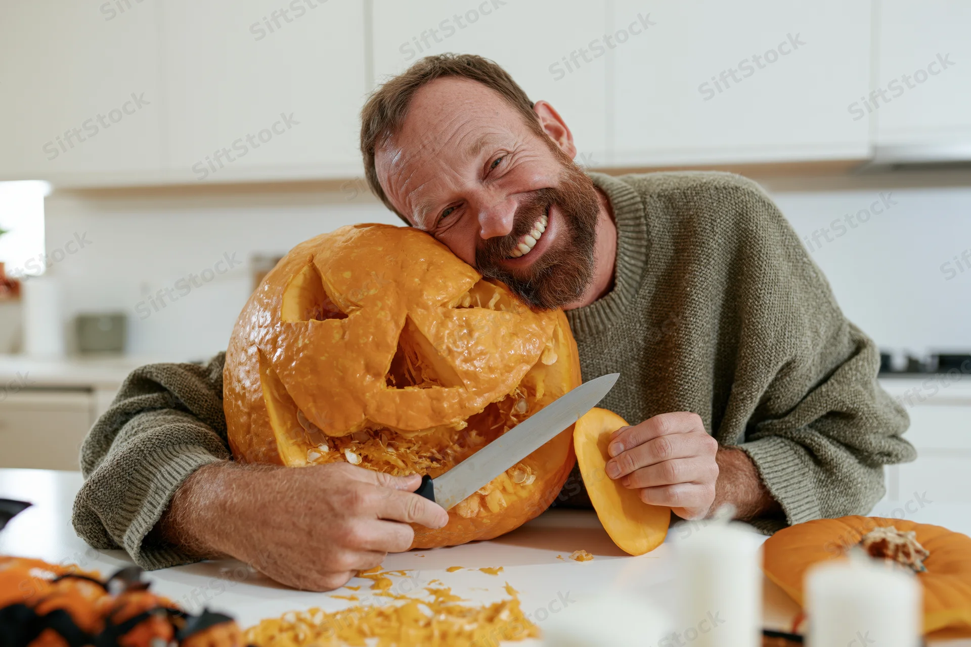 Smiling bearded man leaning cheek-to-cheek against a carved pumpkin while carving it on a kitchen counter