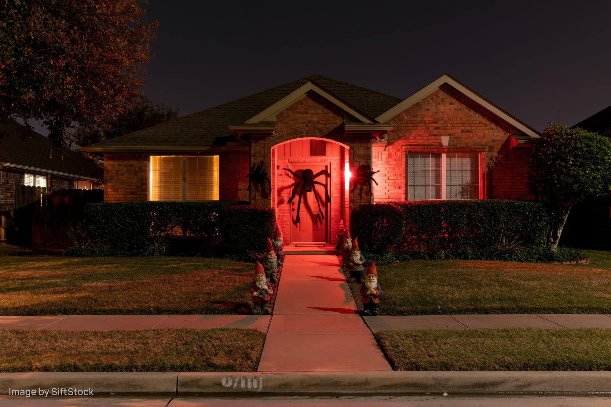 Suburban brick house decorated for halloween at night with red lighting and garden gnomes along the pathway
