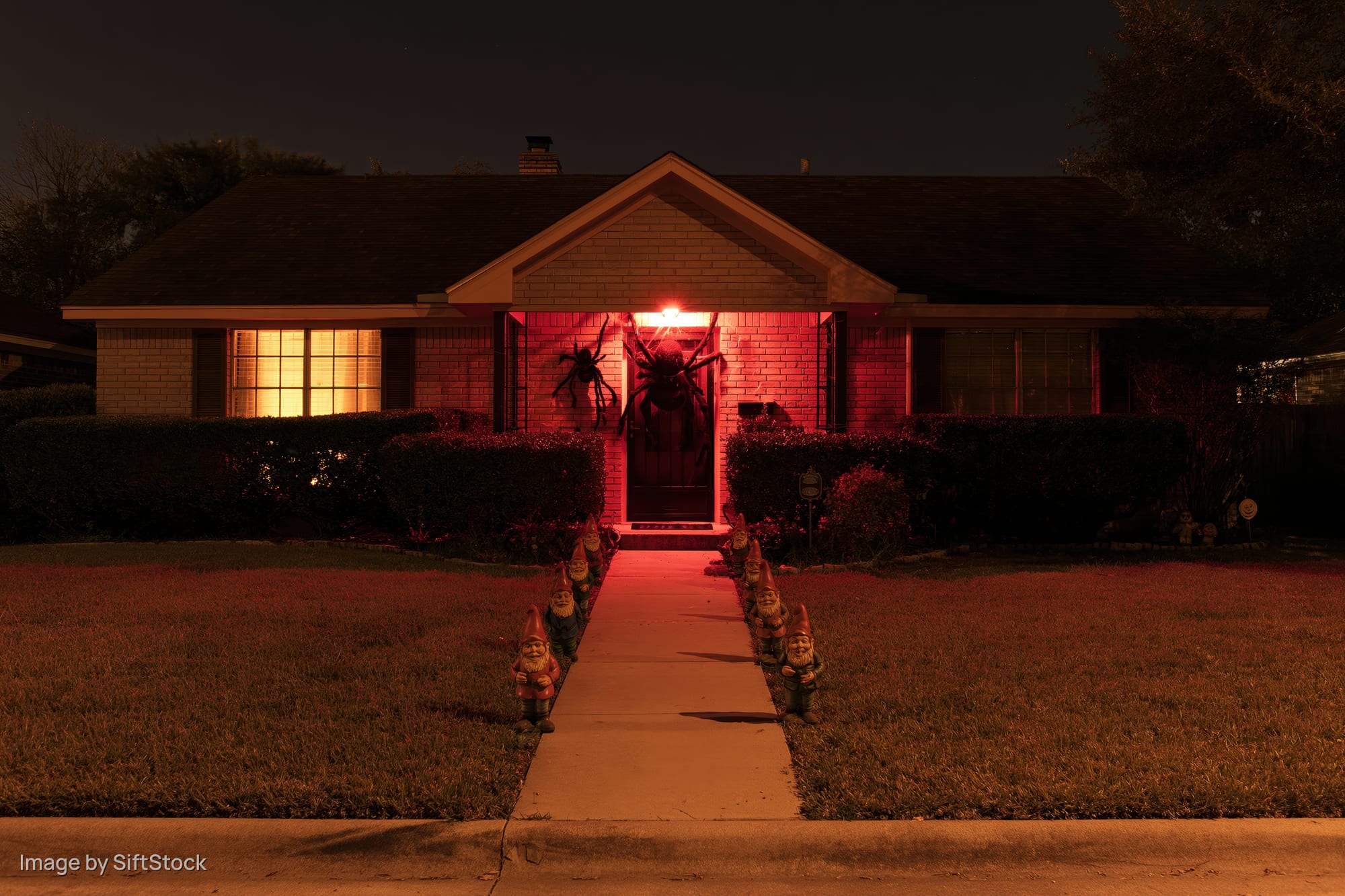 Suburban brick house decorated for halloween at night with red lighting and garden gnomes along the pathway