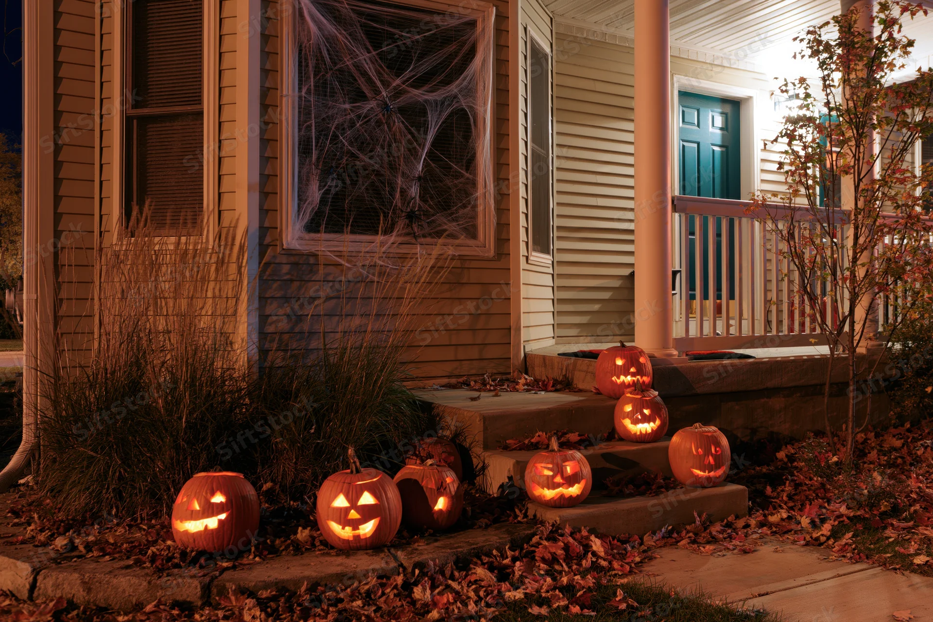 Carved pumpkins glowing on a suburban house porch at night surrounded by autumn leaves and spiderwebs