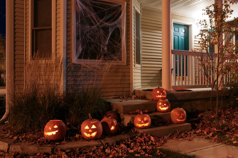 Carved pumpkins glowing on a suburban house porch at night surrounded by autumn leaves and spiderwebs