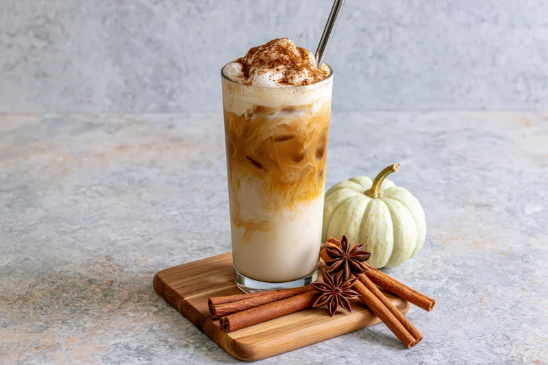 Tall glass of layered iced pumpkin spice latte with caramel swirls, whipped cream and cinnamon topping on a bamboo tray beside a small white pumpkin and cinnamon sticks on a gray stone surface