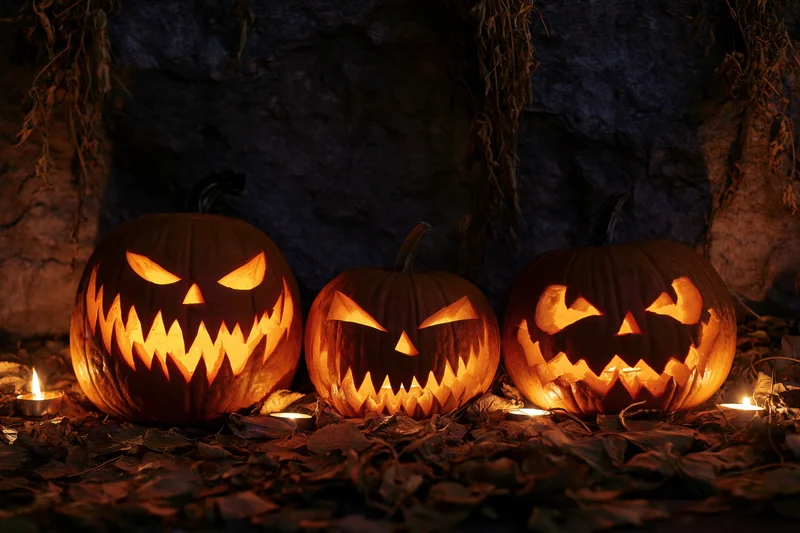 Three carved jack-o-lanterns glowing with candles on a leaf-covered ground at night