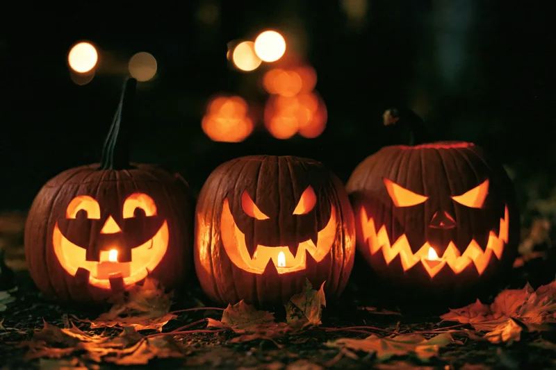 Three carved pumpkins glowing with candles on leaf-covered ground at night