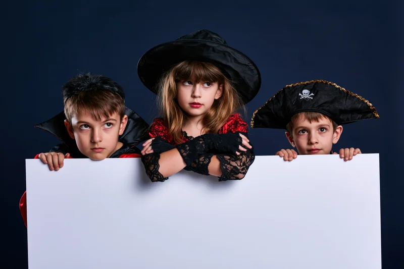 Three children in halloween costumes peeking over a blank white board