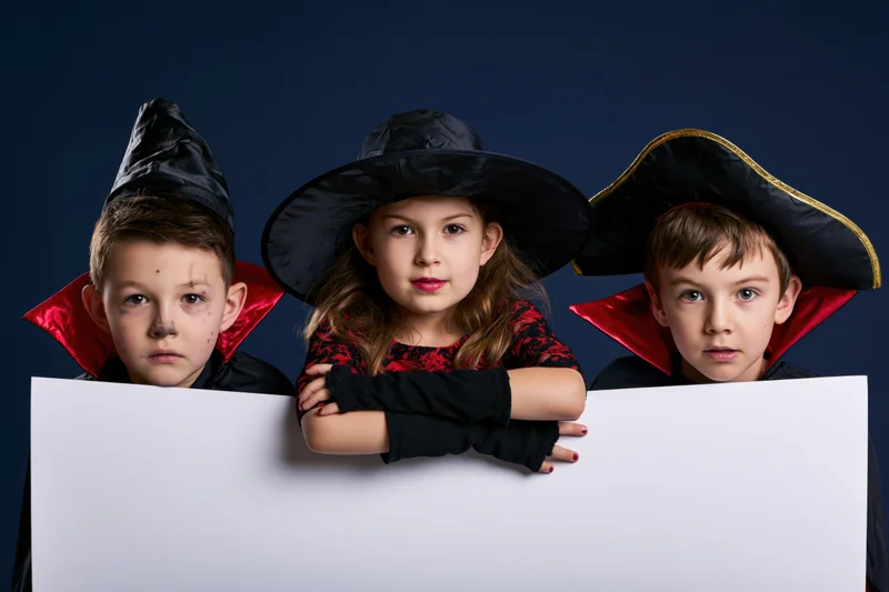 Three children in halloween costumes peeking over a blank white board in studio setting