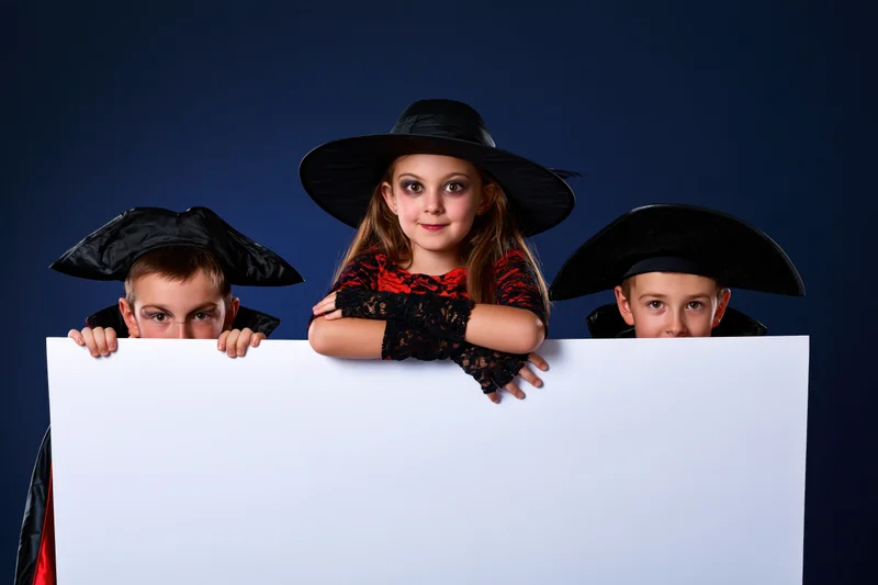 Three children in halloween costumes peeking over a blank white banner in a studio setting