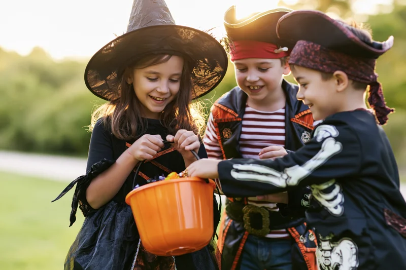 Three children in halloween costumes trick-or-treating outdoors, sharing candy from an orange bucket in a grassy park at sunset