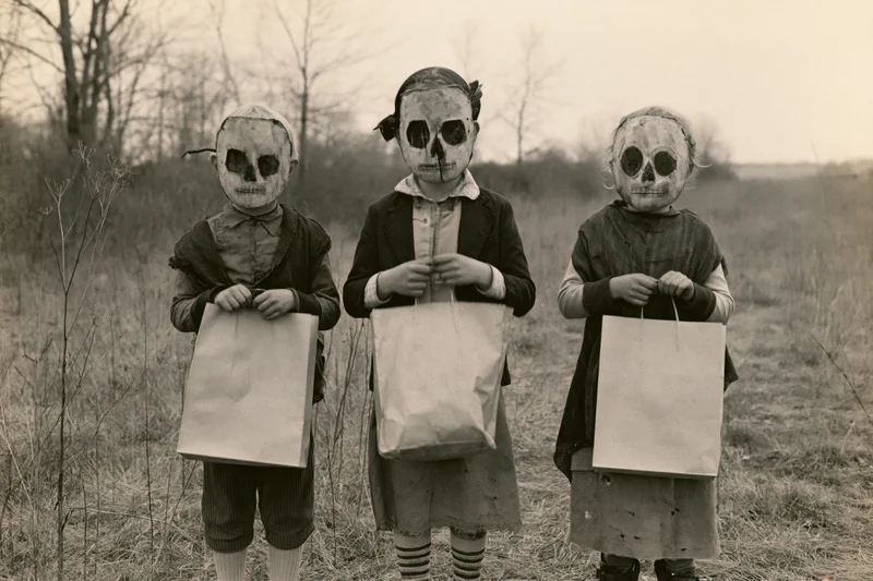 Three children in handmade skull masks holding paper bags in a rural field during halloween