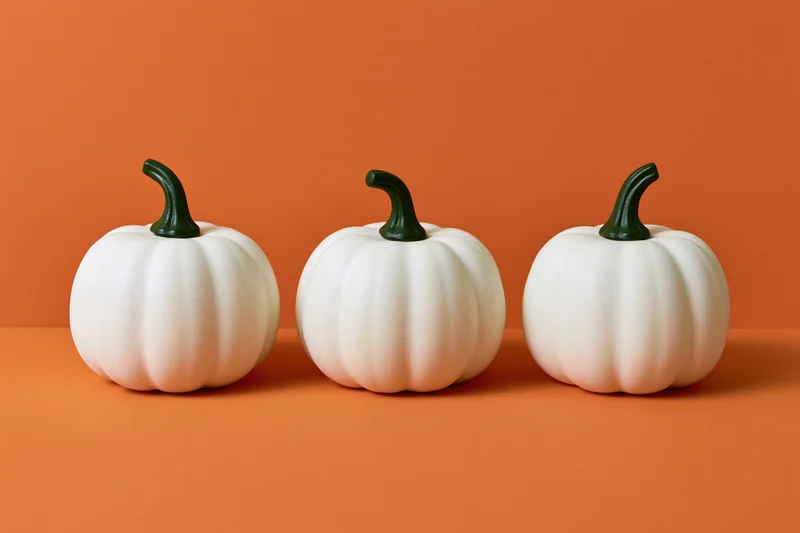 Three white decorative pumpkins with green stems arranged in a row on an orange surface
