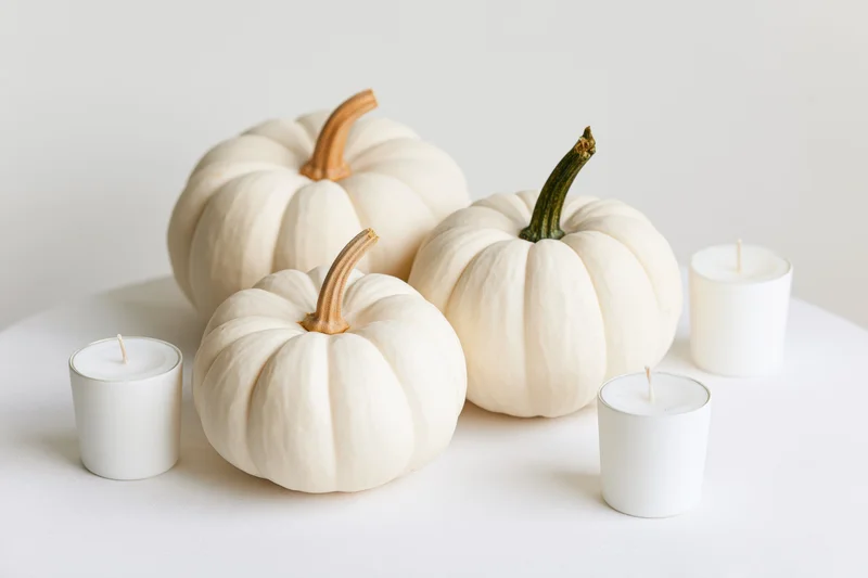 Three white pumpkins with tan and green stems arranged with unlit white candles on a white surface