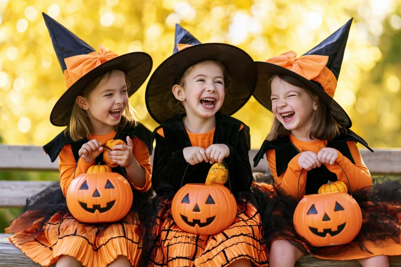 Three young girls in witch costumes laughing together on a bench outdoors
