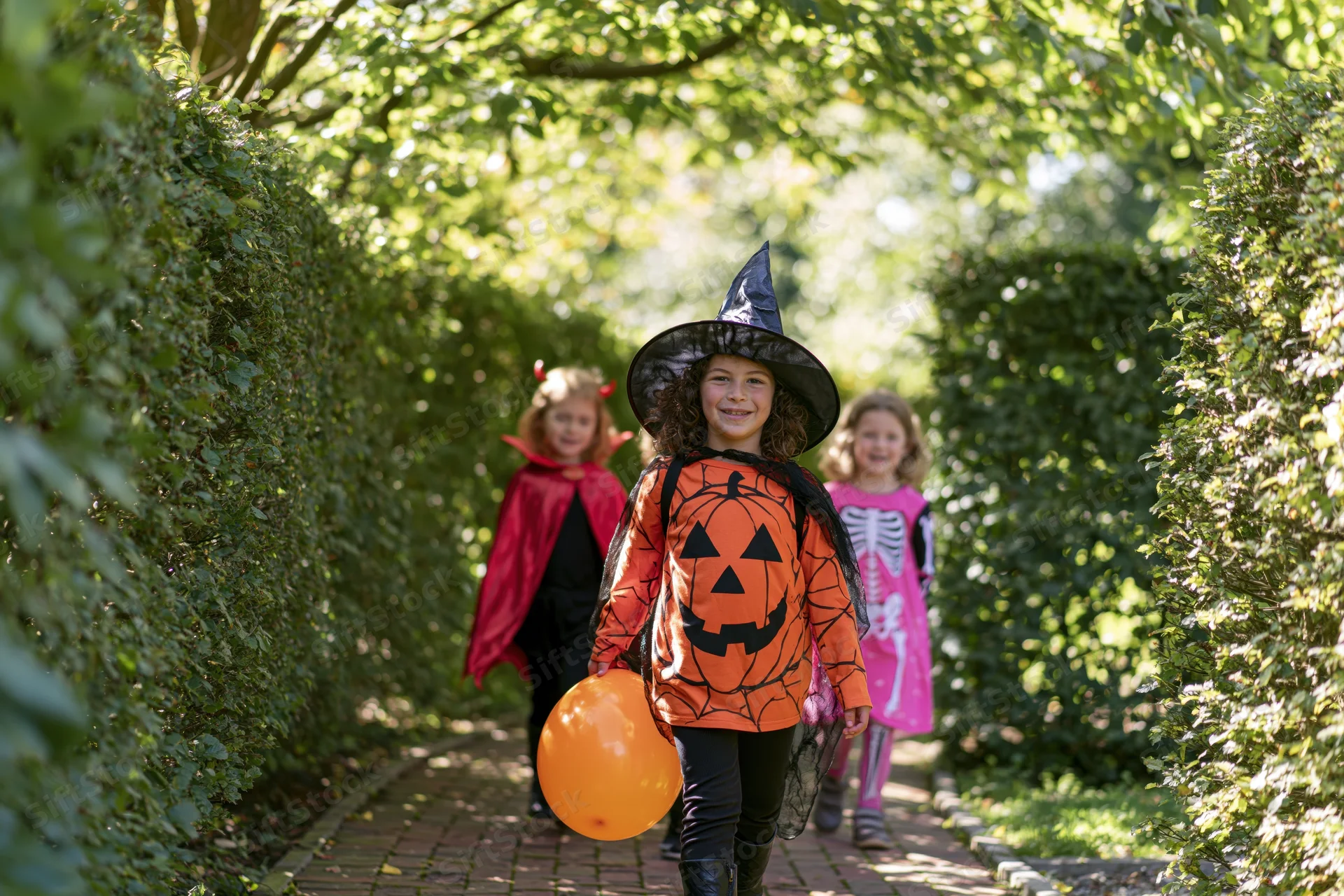 Three children in halloween costumes walking along a garden path toward the camera