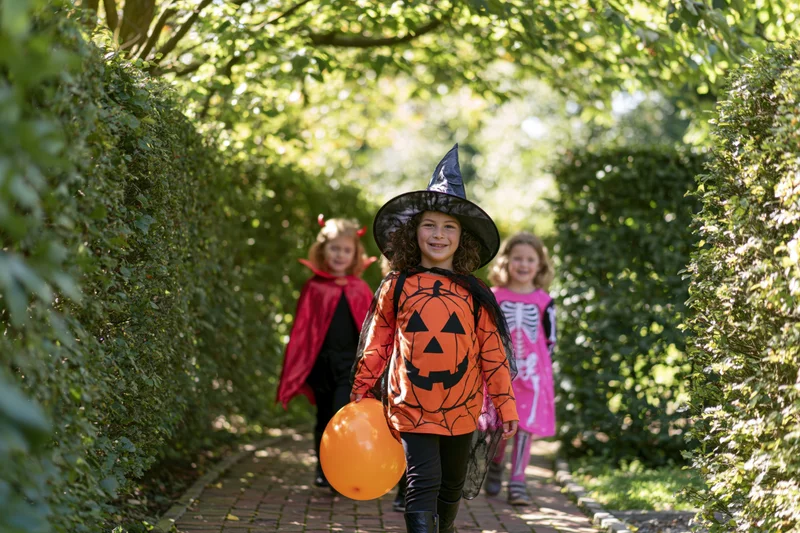 Three children in halloween costumes walking along a garden path toward the camera