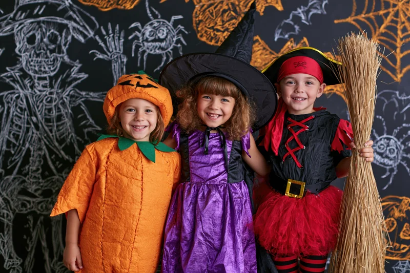 Three young children in halloween costumes posing together indoors smiling in front of a chalkboard with spooky drawings