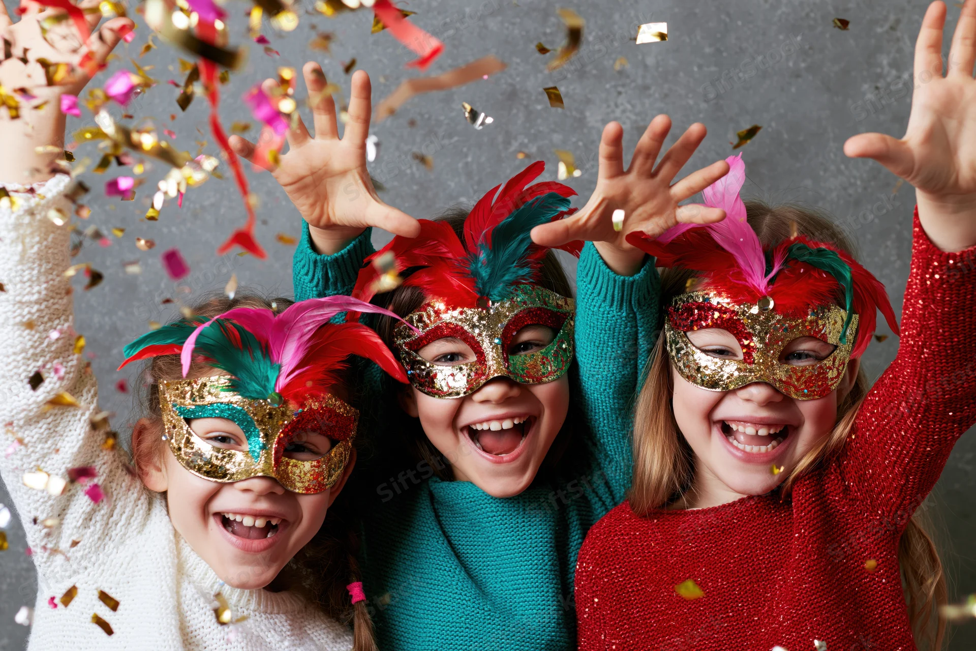 Three young girls wearing colorful venetian masks with feathers, laughing and raising hands amidst falling confetti