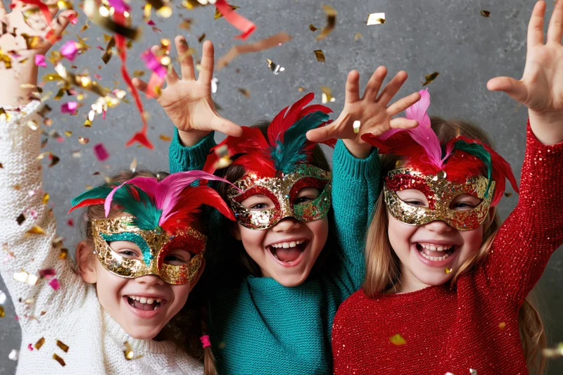 Three young girls wearing colorful venetian masks with feathers, laughing and raising hands amidst falling confetti