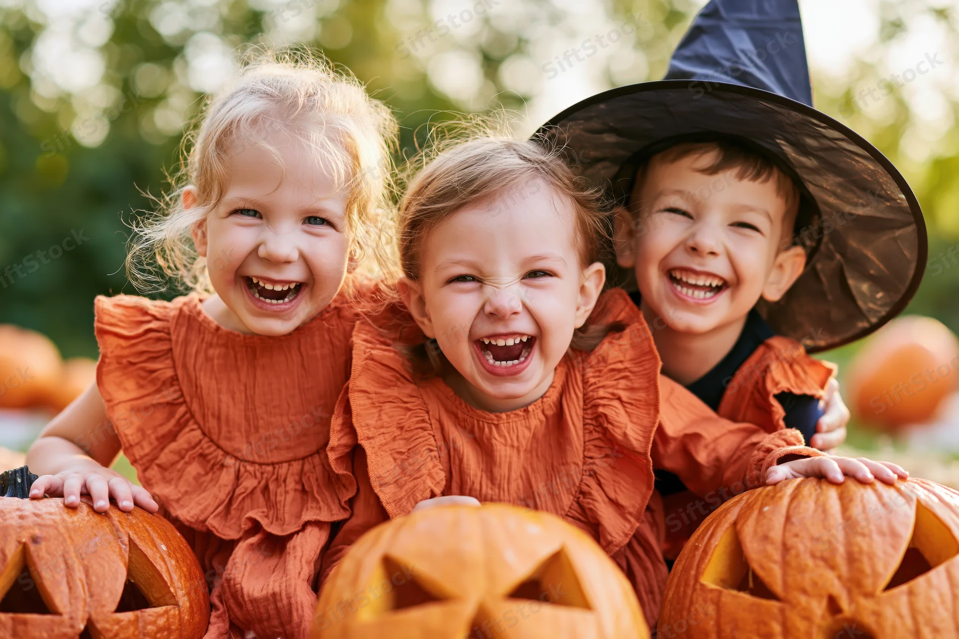 Three toddlers laughing together outdoors with carved pumpkins and halloween costumes