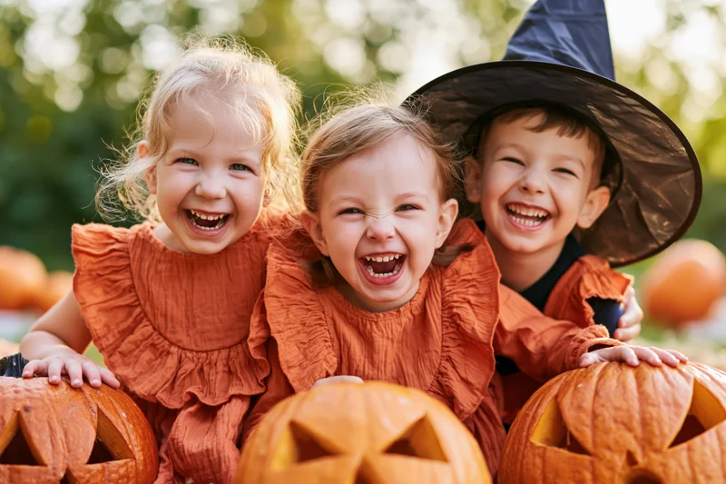 Three toddlers laughing together outdoors with carved pumpkins and halloween costumes