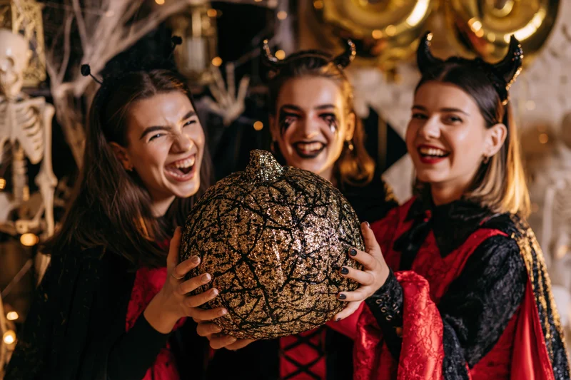 Three young women in halloween costumes laughing while holding a glittery pumpkin at a decorated party