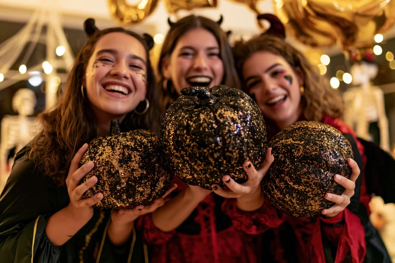 Three young women in halloween costumes laughing while holding glittery black and gold pumpkins indoors