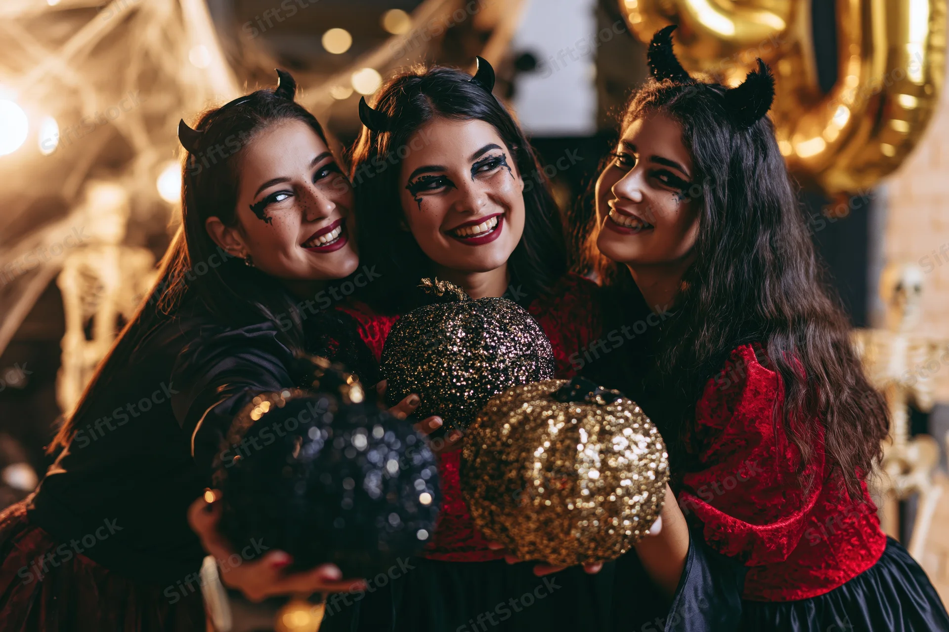 Three young women in devil costumes laughing and holding sequined pumpkins at a halloween party