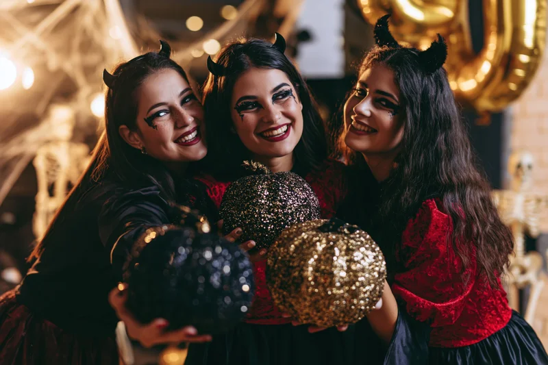 Three young women in devil costumes laughing and holding sequined pumpkins at a halloween party
