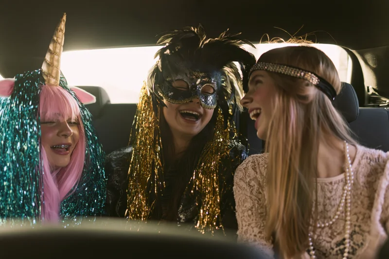 Three young women in party costumes laughing together in a car backseat