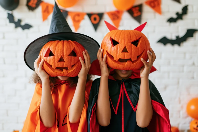 Two children in halloween costumes holding carved pumpkins over their faces in front of a decorated brick wall