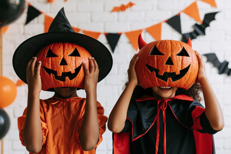 Two children in halloween costumes holding carved pumpkins over their faces indoors