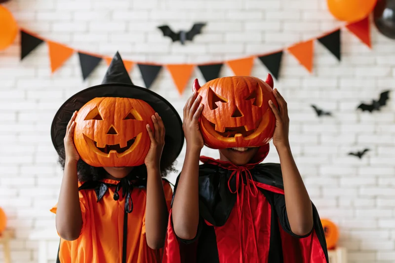 Two children in halloween costumes holding carved pumpkins over their faces indoors