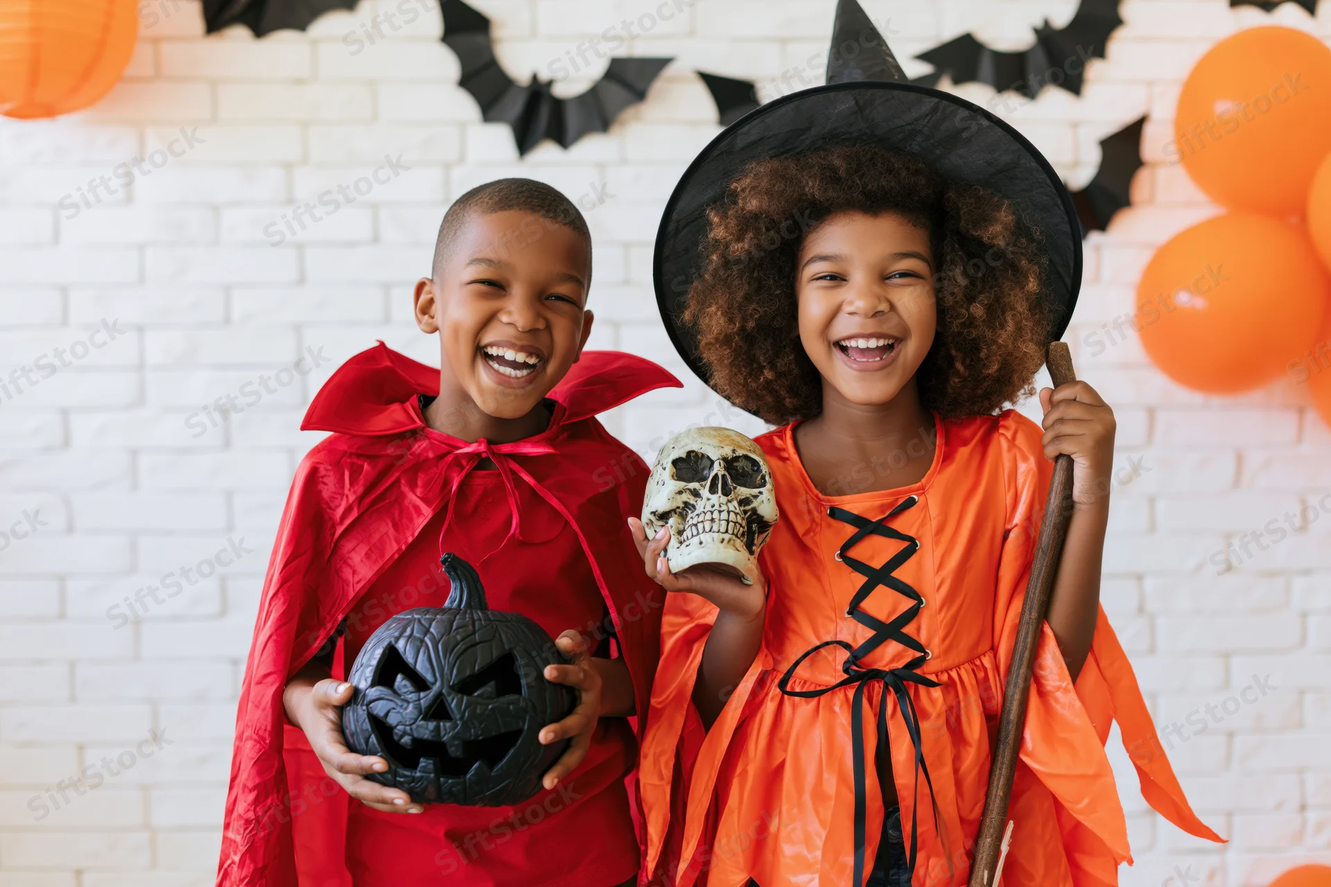 Two black children in halloween costumes laughing together in front of decorated brick wall