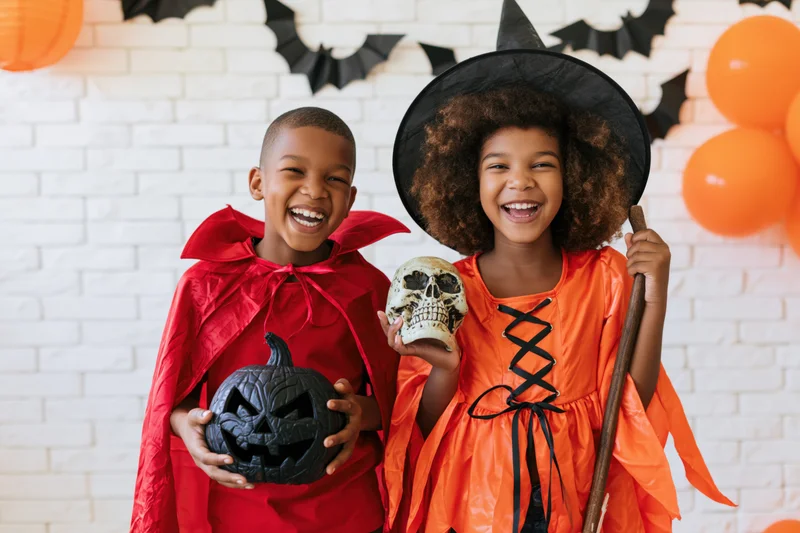 Two black children in halloween costumes laughing together in front of decorated brick wall